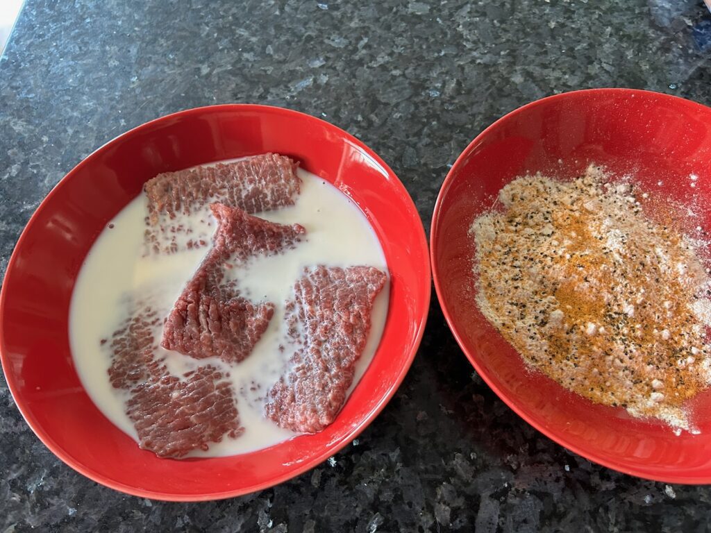 round steak soaking in milk with bowl of seasoned flour nearby