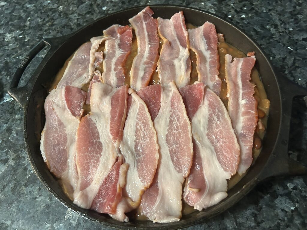 baked beans in cast iron skillet with uncooked bacon on top ready to go in oven
